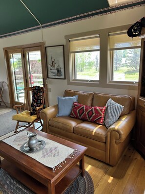 A cozy living room with a brown leather sofa, a wooden coffee table, and a chair with a floral cushion. There are two windows with views of greenery and a door with a glass panel. - Nordic Nook with stars and mountain/color views, cozy and close to town (Durango)