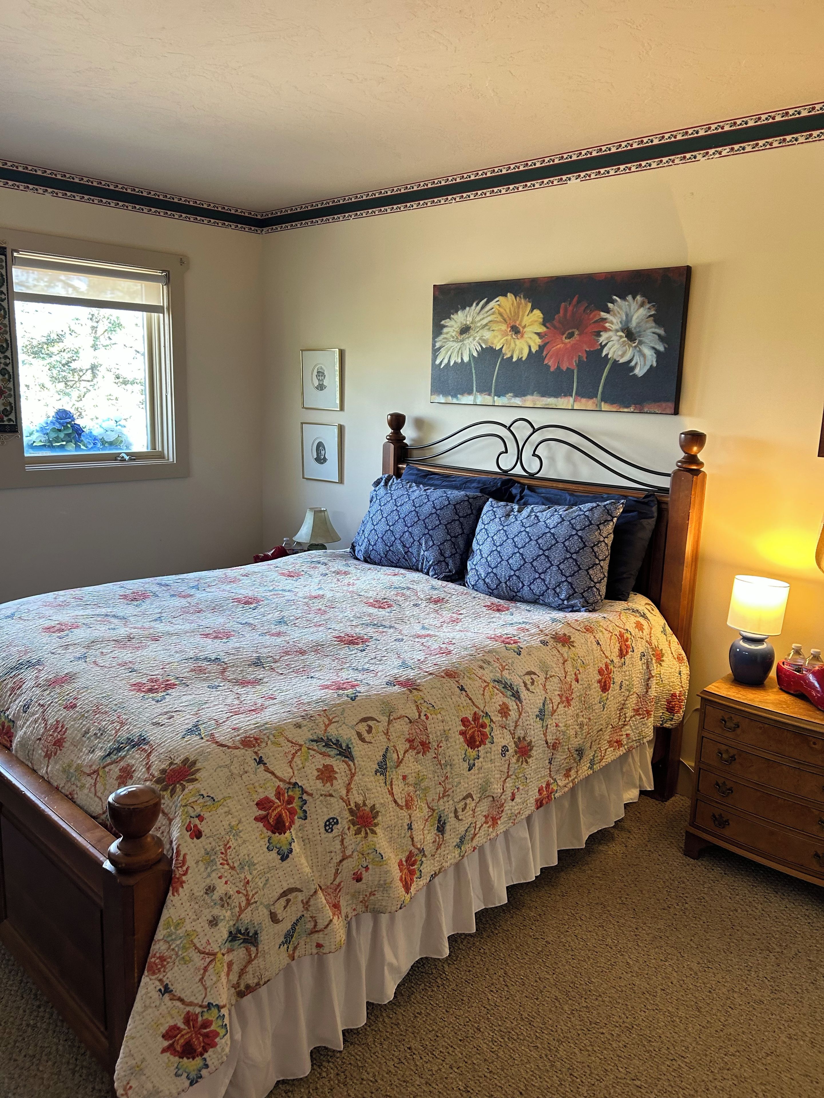 A bedroom with a floral bedspread, a wooden headboard, a nightstand with a lamp, and a window with a view of trees.