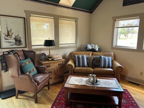 A living room with a brown leather armchair, a brown sofa with patterned cushions, a wooden side table with a lamp, and a coffee table with a metal pitcher.