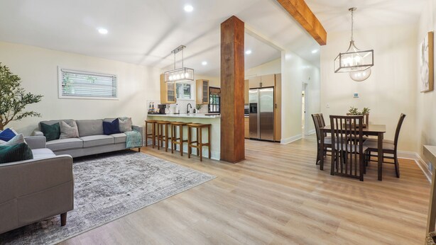 A spacious living area with a sofa, coffee table, and a dining table with chairs. There is a kitchen area with modern appliances and a wooden ceiling beam.