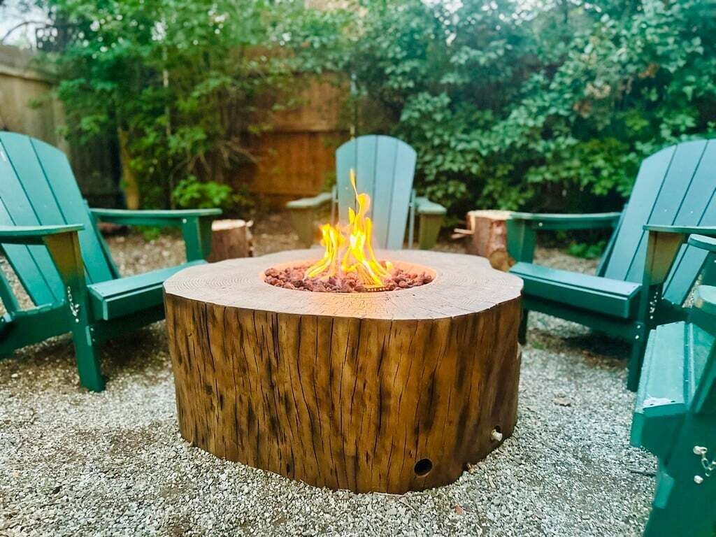 Ein runder, hölzerner Feuerkorb mit brennendem Feuer in der Mitte, umgeben von grünen Adirondack-Stühlen.