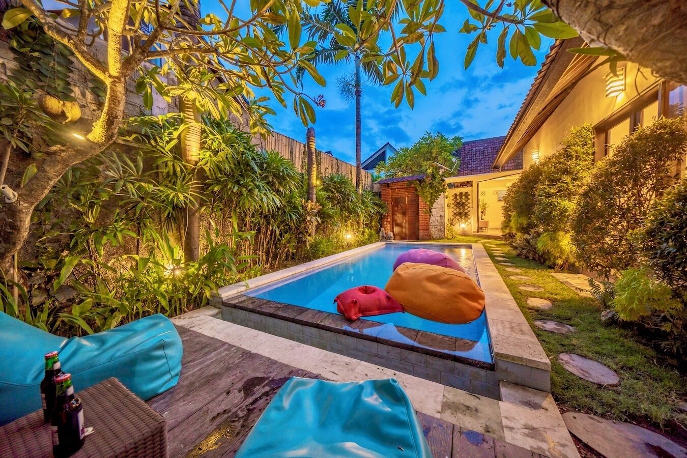 A swimming pool with colorful bean bags, surrounded by lush greenery and a well-lit building in the background.