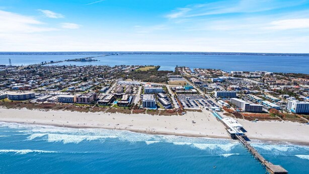 Vue aérienne d’une ville côtière avec une plage, une jetée et divers bâtiments, notamment des hôtels et des zones résidentielles.