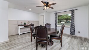A dining area with a round table, six chairs, a ceiling fan, and a window with patterned curtains.