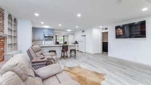 A spacious living area with a sofa set, a coffee table, and a rug. A kitchen with white cabinets and a dining area with a table and chairs is visible in the background.