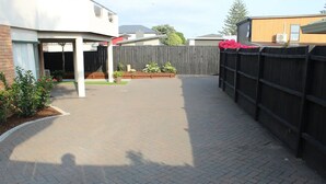 A paved backyard with a wooden fence, a covered patio area, and potted plants.