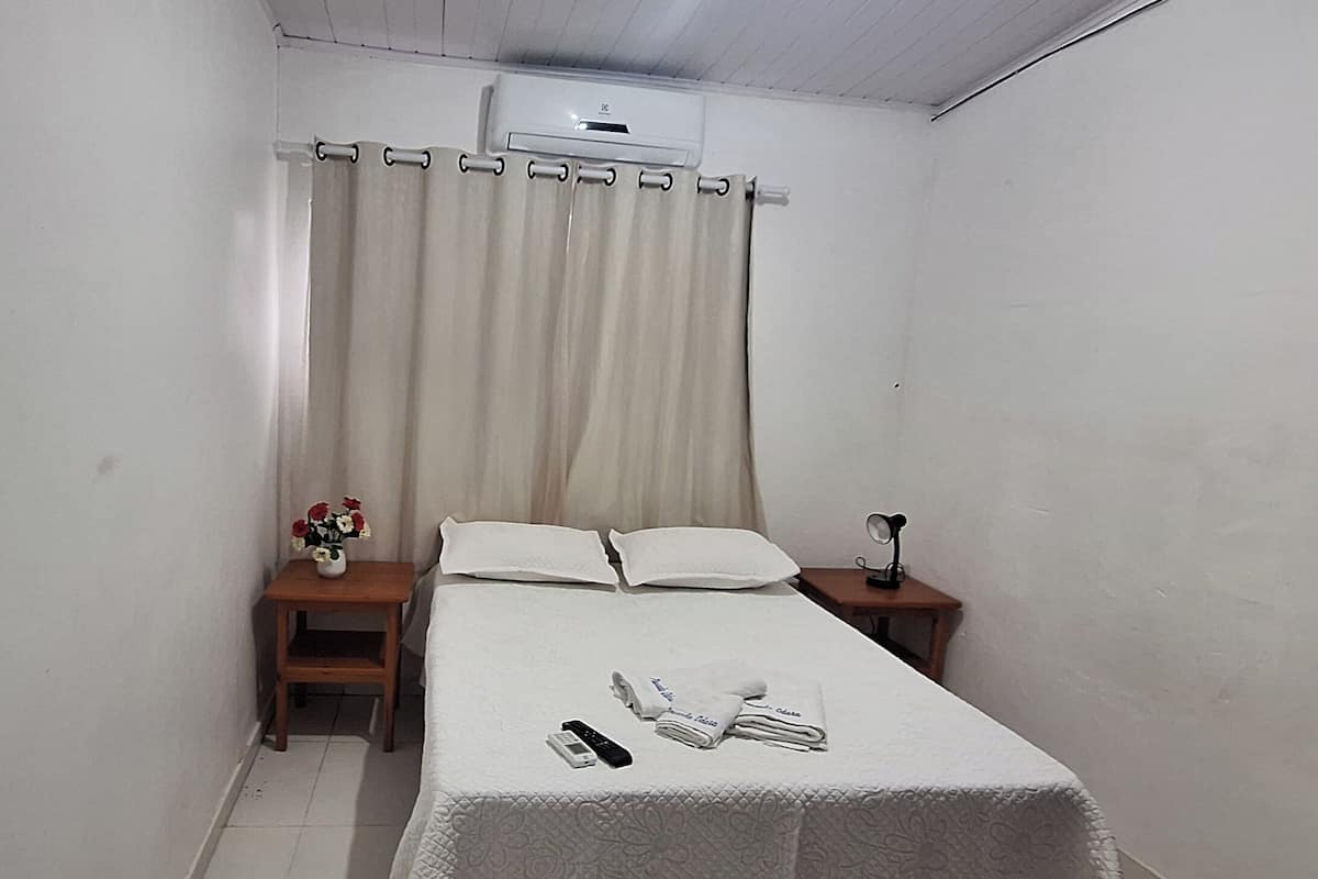 A small, neatly arranged bedroom with a single bed, a bedside table with a lamp, and a wall-mounted air conditioner.