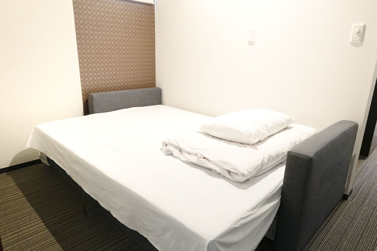 Un lit simple avec un matelas et des oreillers blancs, une tête de lit grise et un banc recouvert d’un tissu gris.