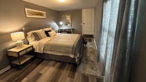A bedroom with a bed, bedside table, lamp, dresser, mirror, and a window with curtains. - corporate comfort suits  (Pocahontas)