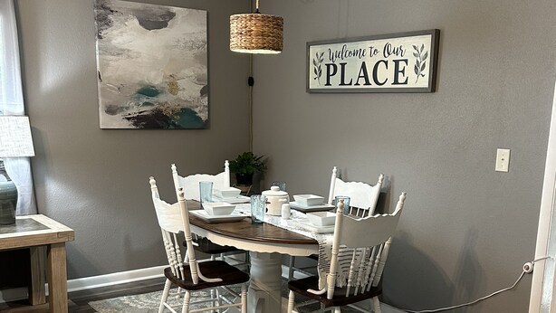 A dining area with a wooden table, white chairs, and a rug. There is a painting on the wall and a sign that says "Welcome to Our Place". - corporate comfort suits  (Pocahontas)
