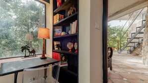 A room with a bookshelf, a desk, and a chair. There is a window with a view of trees and a stone staircase.