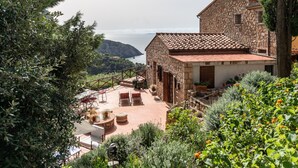 A stone building with a terrace, surrounded by greenery and overlooking a coastal landscape.