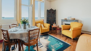 Un comedor con una mesa dispuesta para cuatro personas, un sillón amarillo, una chimenea y vistas al mar a través de amplios ventanales.