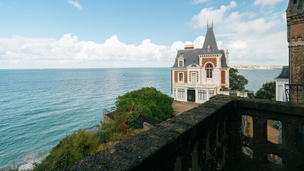 Un edificio histórico con una torre destacada, con vista a un mar tranquilo y a una costa distante.