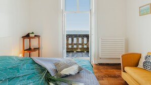 Un dormitorio con cama, una mesita con una planta, un radiador y vista al mar a través de una puerta abierta.