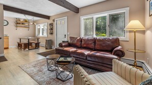 Ein Wohnzimmer mit einem braunen Ledersofa, einem gestreiften Sessel und einem Holztisch. Eine Lampe, ein Teppich und ein Fenster mit Blick auf Bäume sind ebenfalls vorhanden.