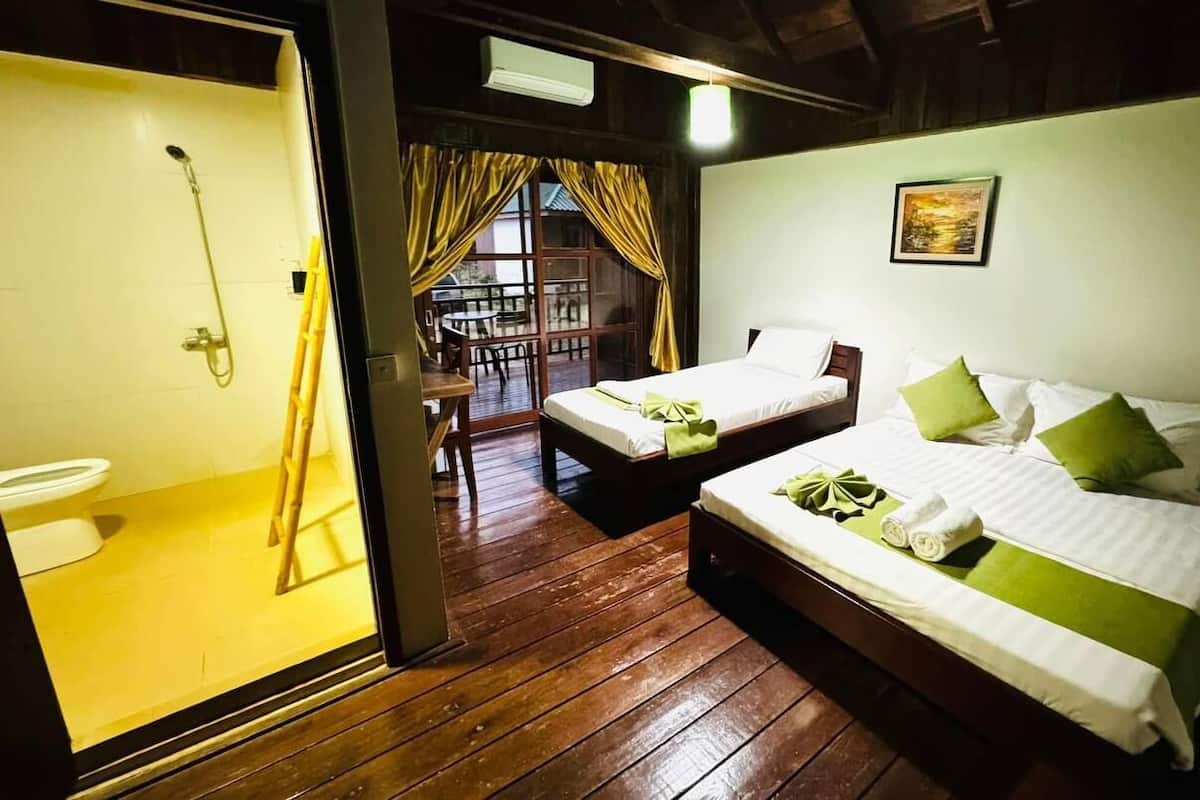 A room with two beds, a wooden floor, a bathroom with a shower, and a balcony with a table and chairs.