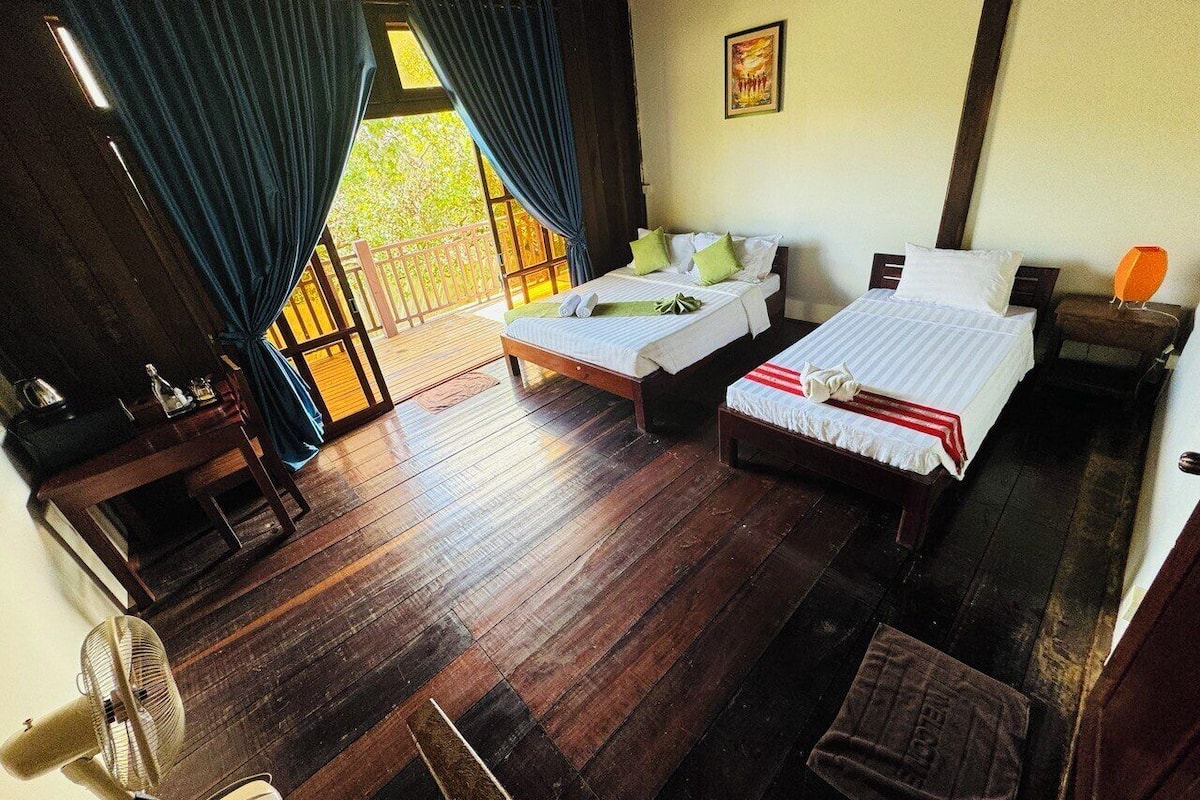 A room with two beds, a wooden floor, a fan, a table with a lamp, and a view of greenery outside.