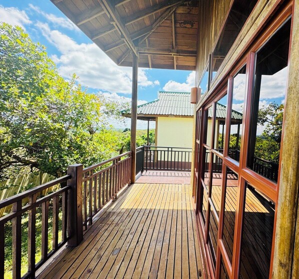 A wooden deck with a railing, large windows, and a view of greenery and a building in the background. - RAtanakiri PAradise Resort  (Ban Lung)