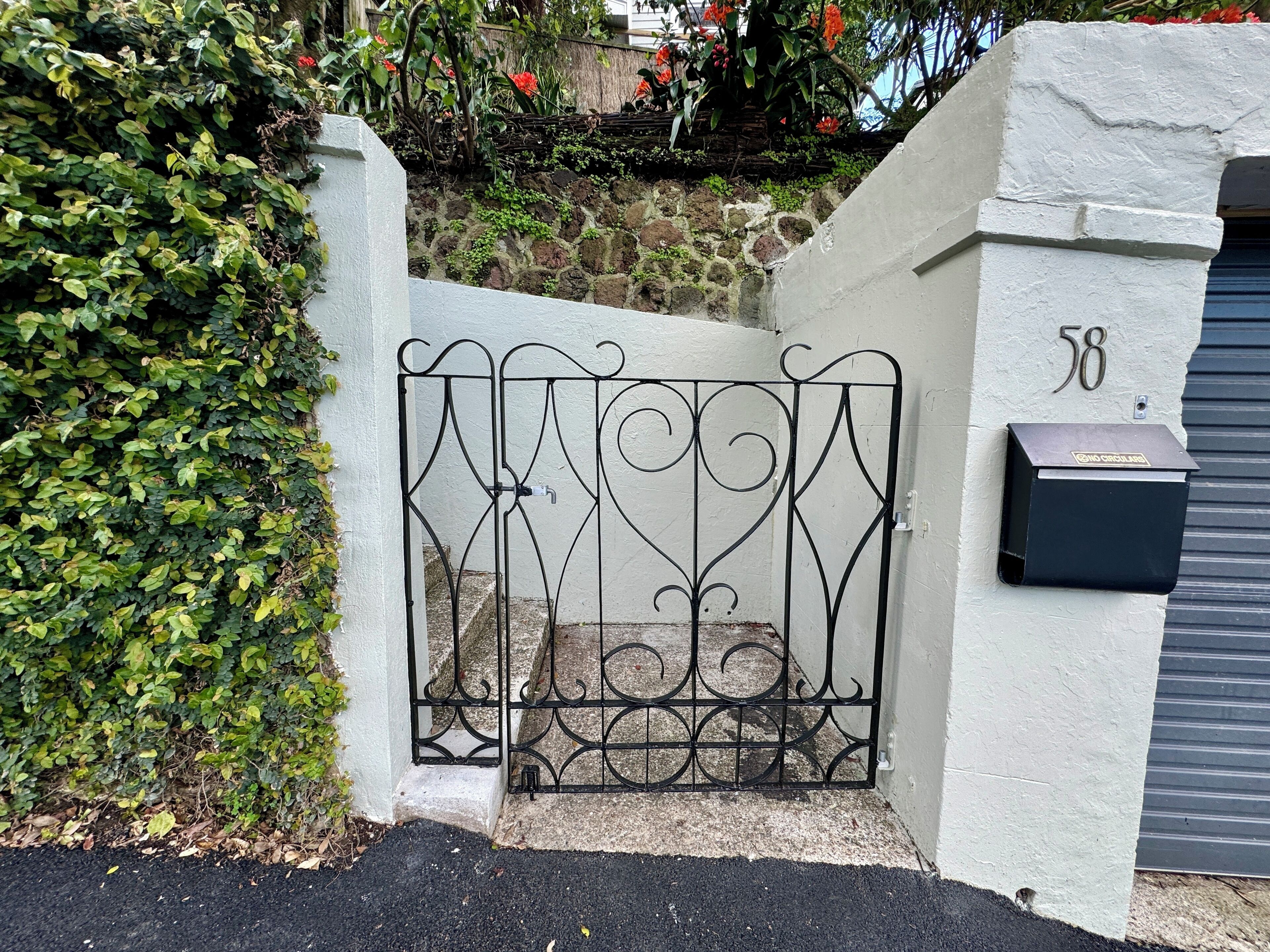 Une entrée fermée par un portail, avec une boîte aux lettres, un mur en pierre et une haie.