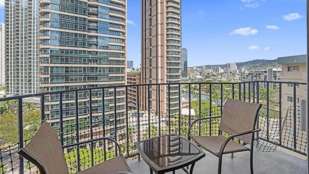 Un balcon avec une table en verre et deux chaises, donnant sur un paysage urbain avec de hauts immeubles et une rivière.