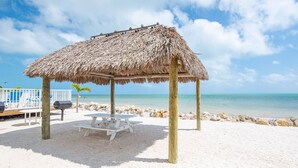Une plage avec un pavillon à toit de chaume, une table de pique-nique blanche et des chaises longues.