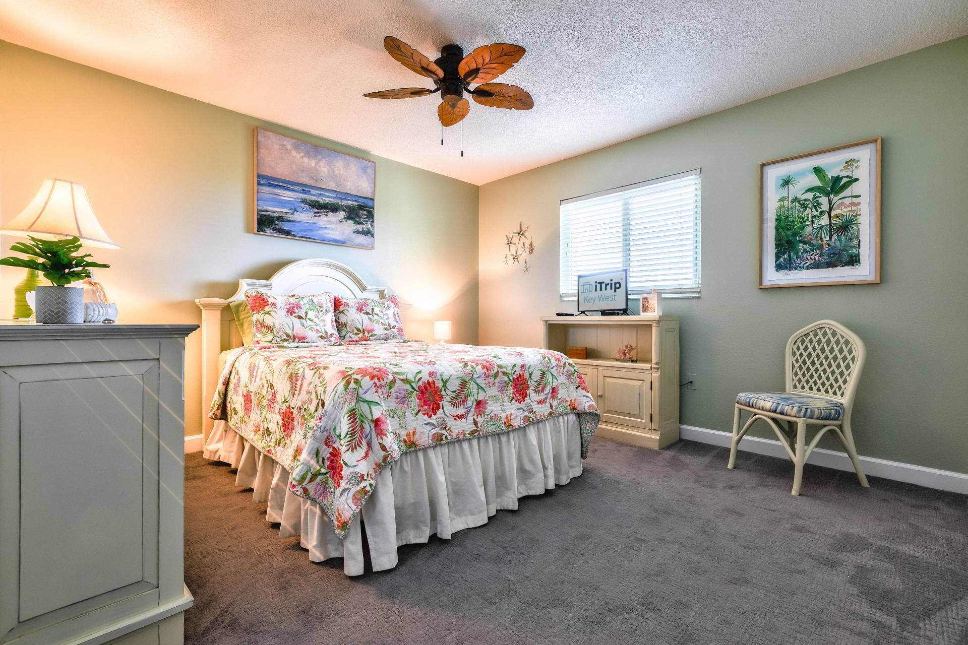 A bedroom with a bed, a chair, a desk, a ceiling fan, and artwork on the walls.