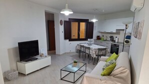 Ein modernes Wohnzimmer mit Flachbildfernseher, einer Couch mit grünen Kissen, einem Esstisch und einer Küchenzeile mit Schränken und Herd.