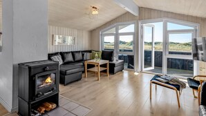 A cozy living room with a wood-burning stove, a black sofa, and a wooden coffee table. Large windows offer a view of the outdoors. - "Jaana" - 10m from the sea by Interhome (Blåvand)