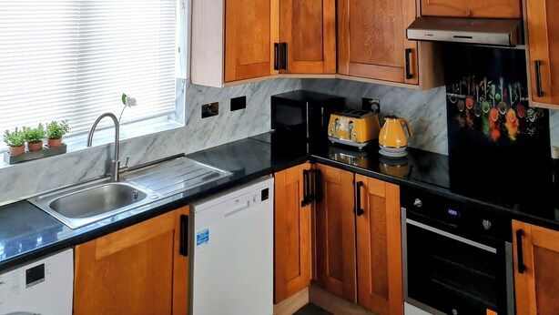 Una cocina con armarios de madera, encimera negra, pileta de acero inoxidable, lavavajillas blanco, refrigerador negro con decoraciones de frutas coloridas y tres lámparas colgantes.