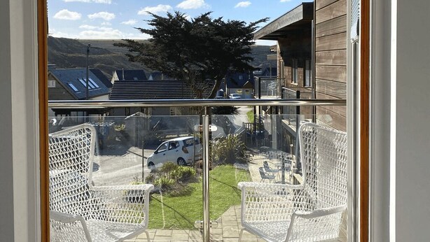 A balcony with two white wicker chairs, a glass railing, and a view of a car and buildings in the background. - Porthtowan Coastal Escape with minutes away from the Beach (Truro)