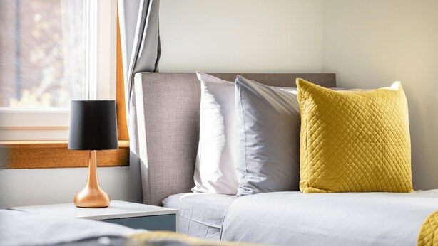 Una cama bien hecha con un cabecero gris, un cojín acolchado amarillo y una lámpara negra sobre una mesita de noche.
