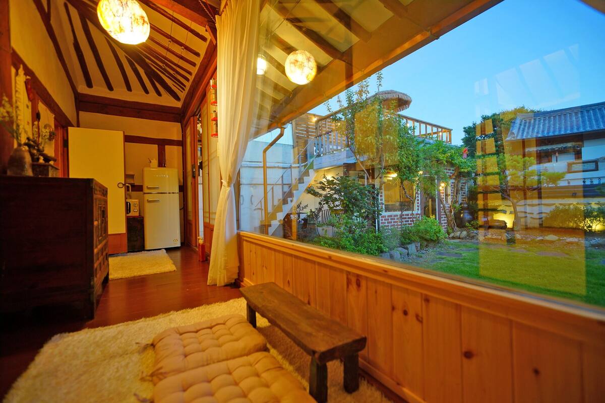A cozy room with a wooden bench, a refrigerator, and a view of a garden and buildings outside.