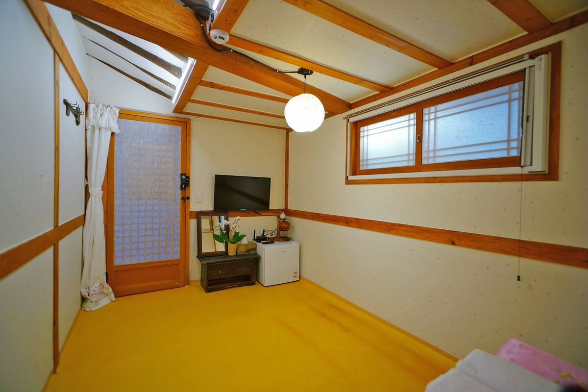 A small room with a yellow floor, wooden beams, a television, a small table with a plant, and a window with blinds.