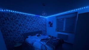 Ein Schlafzimmer mit einem gemusterten Wandteppich, einem Bett mit einer gemusterten Tagesdecke, einem Nachttisch, einem Fenster mit Jalousien und blauen LED-Leuchten an der Decke.