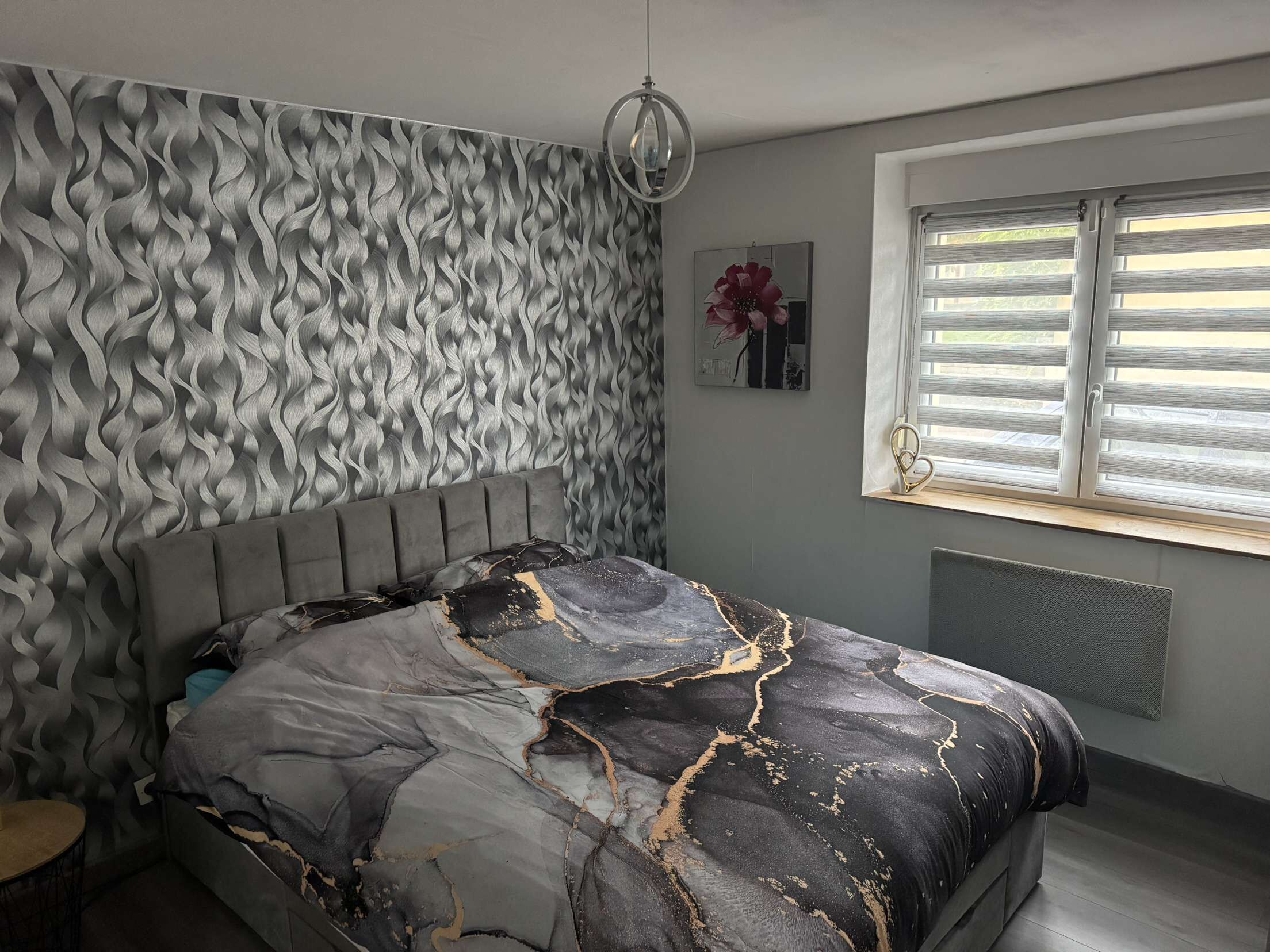 Un dormitorio con papel tapiz estampado, una cama con cubierta que imita mármol, una mesita de noche, una ventana con persianas y una obra de arte colgada en la pared.