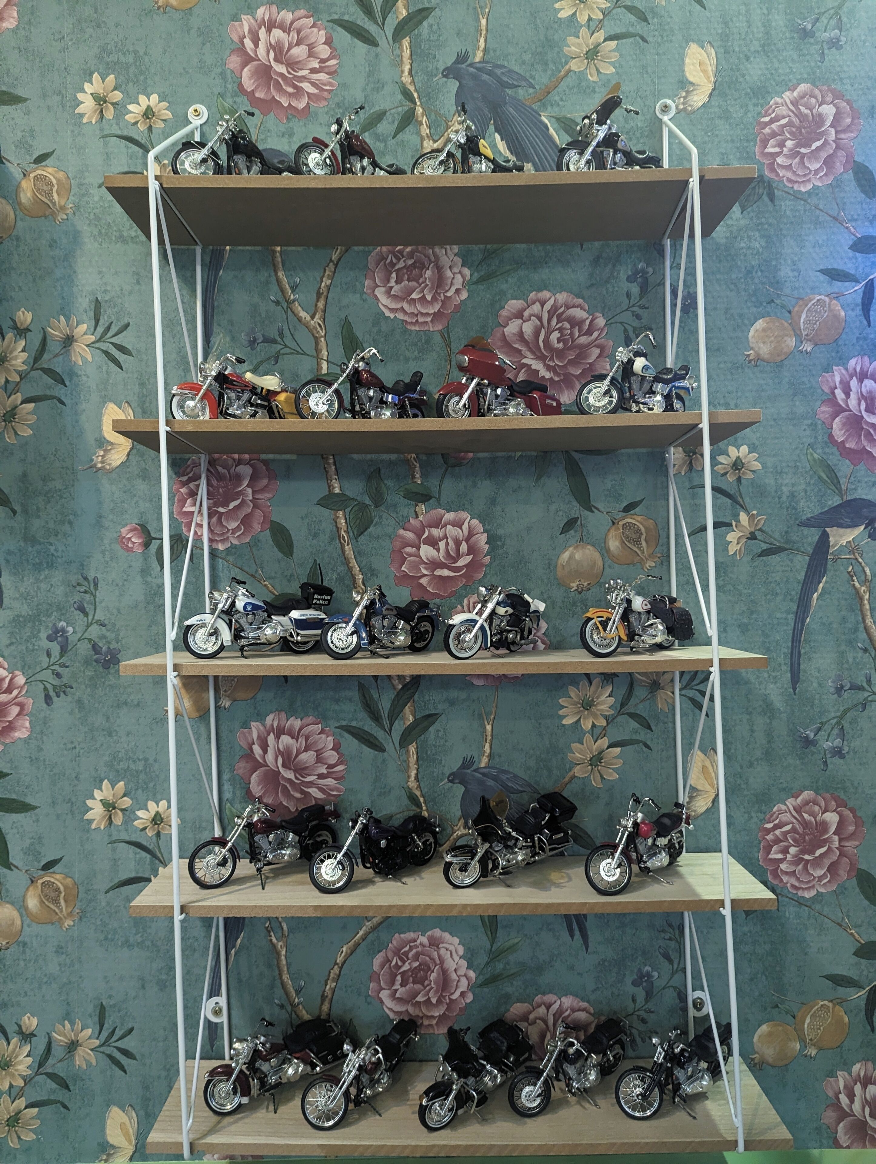 Une étagère à plusieurs niveaux, où sont exposés différents modèles de motos, le tout sur un fond de papier peint à motifs floraux.