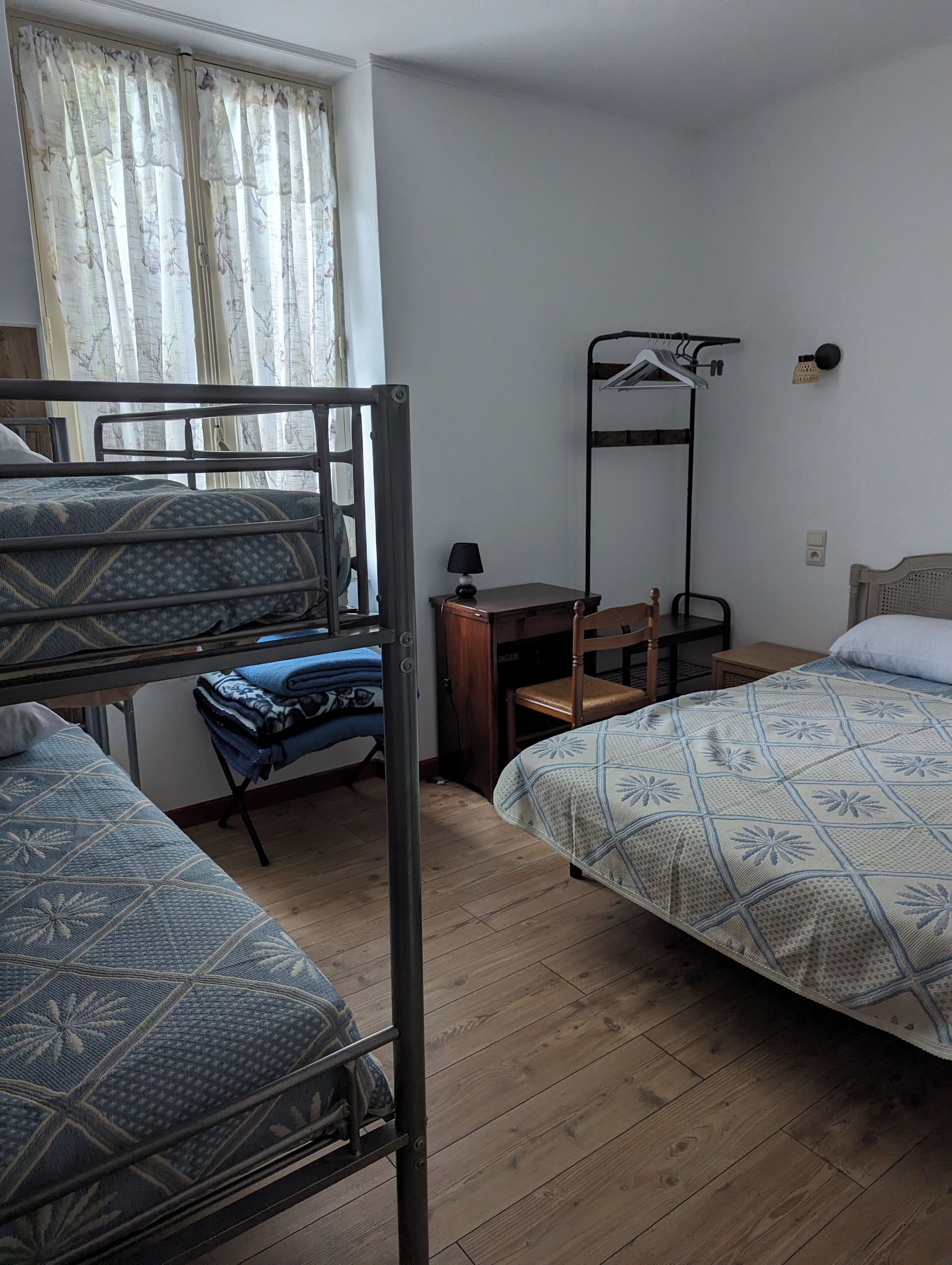 A dormitory room with bunk beds, a desk, a chair, and a hanging clothes rack.