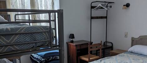 A dormitory room with bunk beds, a desk, a chair, and a hanging clothes rack.