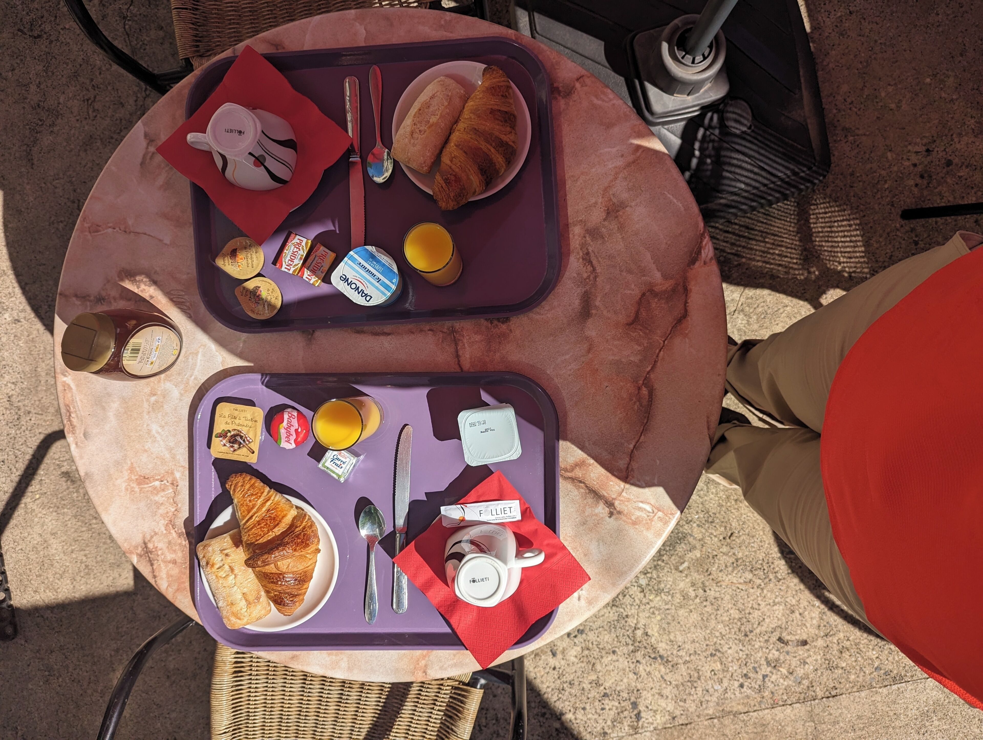 Ein runder Tisch mit zwei Tabletts mit Frühstücksartikeln, darunter Croissants, Brot, eine Saftflasche, eine Tasse und eine kleine Dose Brotaufstriche.