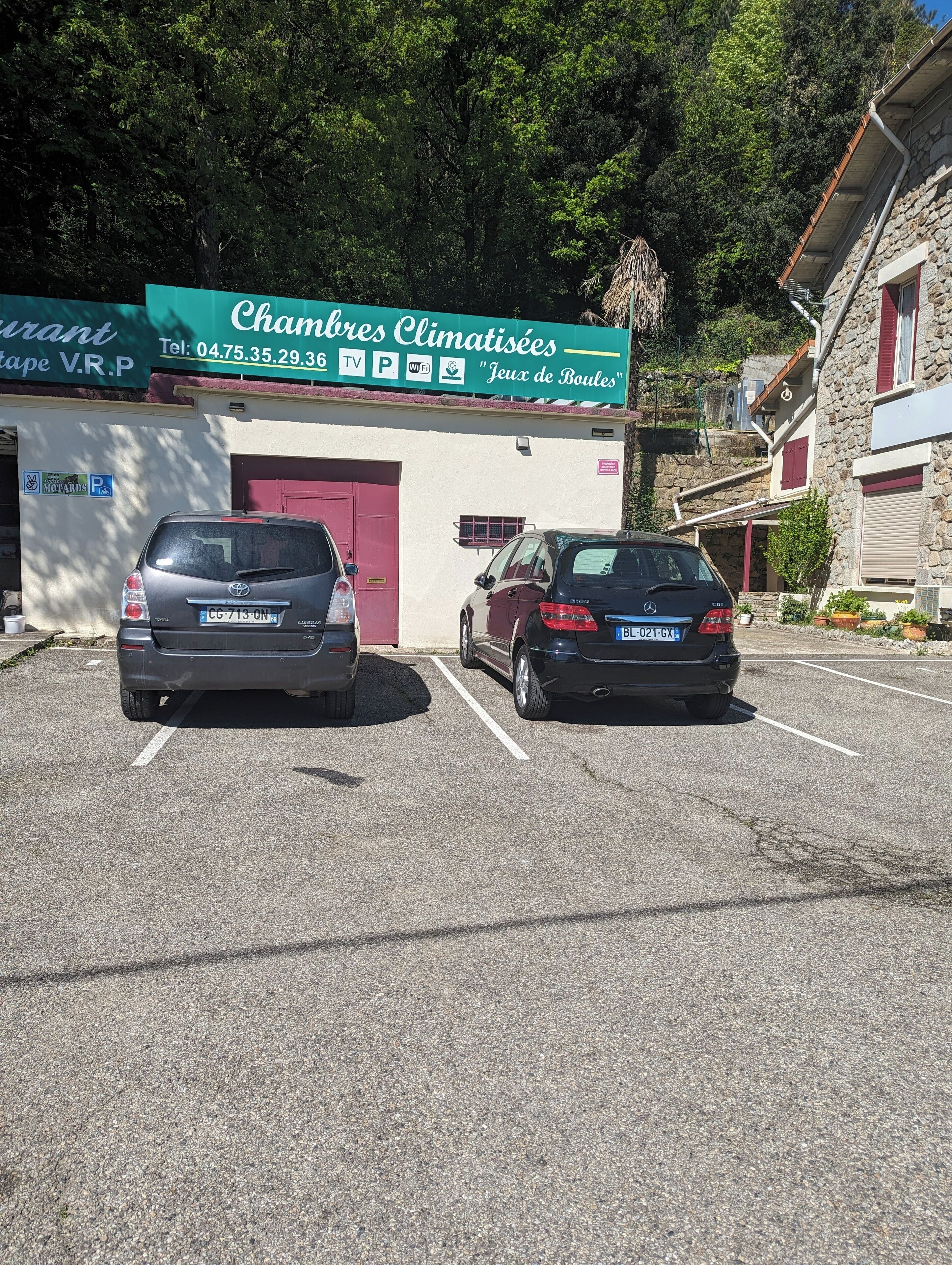 Un estacionamiento con dos autos estacionados frente a un edificio que tiene un letrero con la frase 'Chambres Climatisees' y 'Jeux de Boules'.