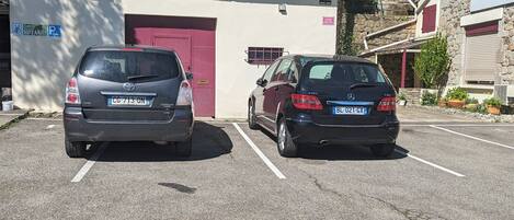 A parking lot with two cars parked in front of a building with a sign for 'Chambres Climatisees' and 'Jeux de Boules'.