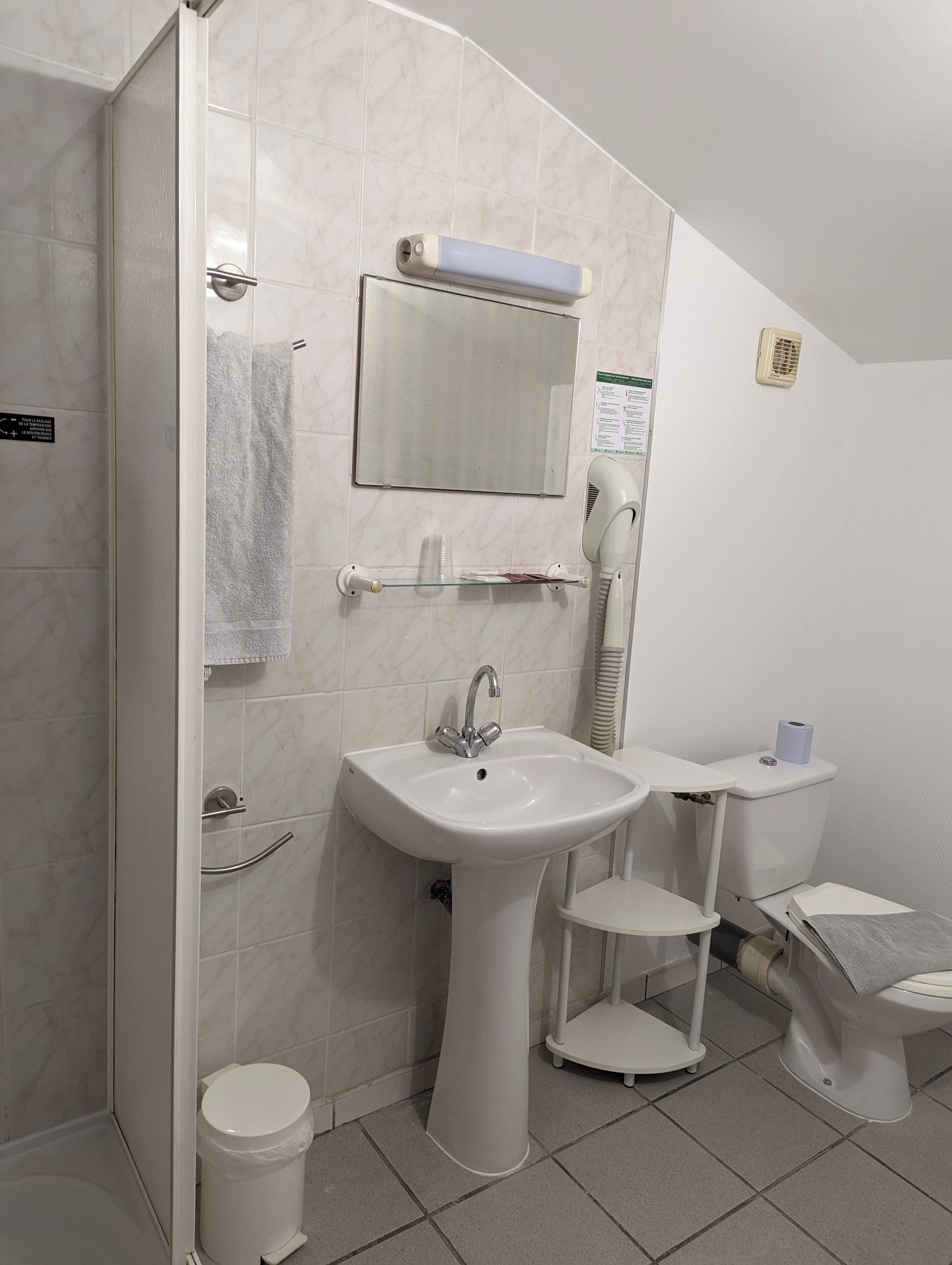 A bathroom with a white sink, mirror, and toilet. There is a towel rack, a hairdryer, and a small shelf.