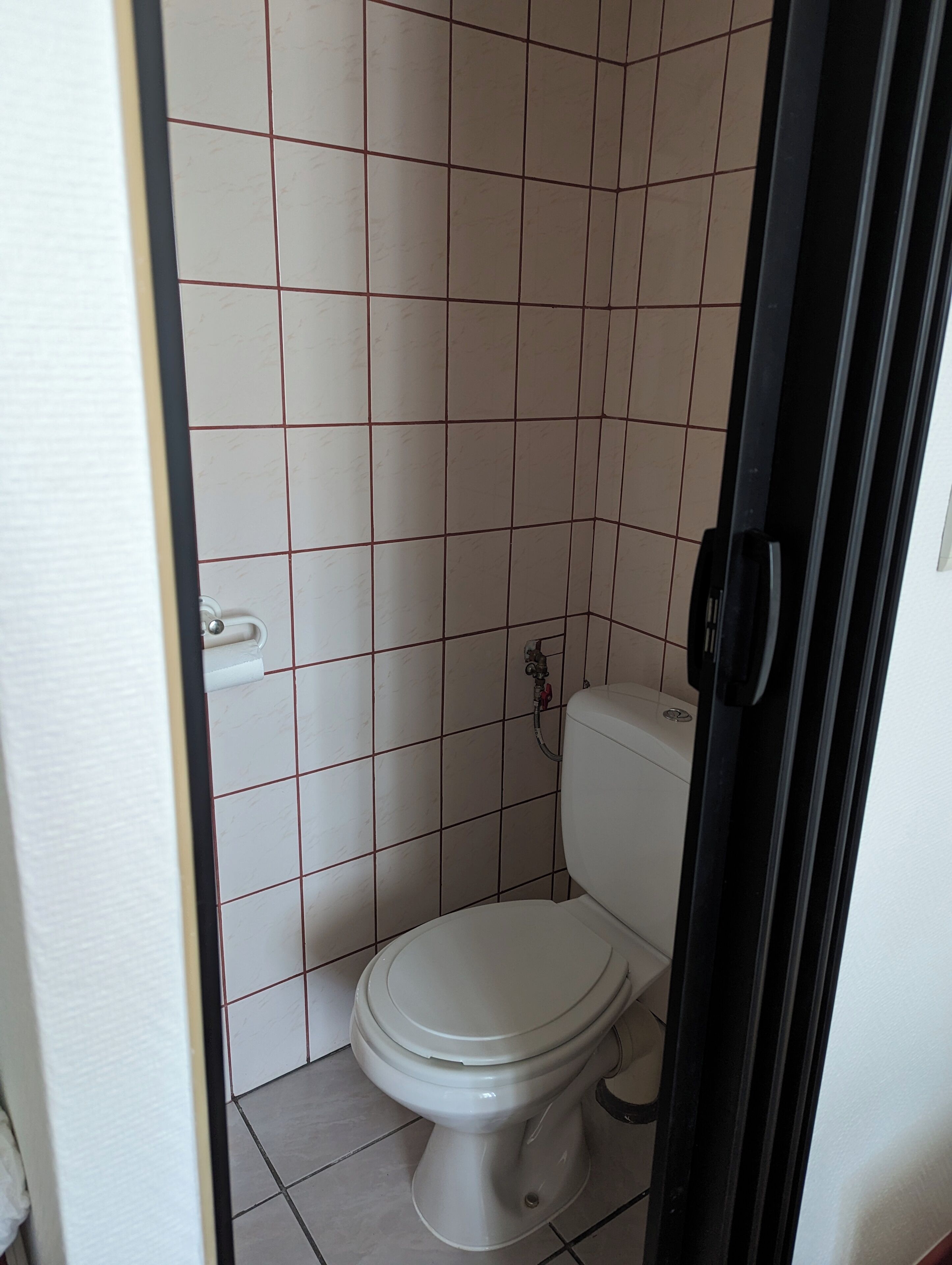 Une petite salle de bain carrelée, avec des toilettes, un robinet d’eau apparent et un cadre de porte.