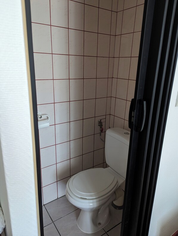 A small, tiled bathroom with a toilet, a visible water valve, and a door frame. - AUBERGE DES PINS (Aubenas)