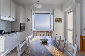 Une cuisine avec des meubles blancs, un fourneau et une table à manger avec des chaises. Une fenêtre offre une vue sur la mer.