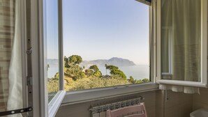 Quarto com uma grande janela com vista para a paisagem costeira, com árvores e colinas.