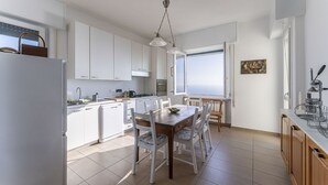 Uma cozinha moderna com armários brancos, uma mesa de jantar com cadeiras e uma janela grande com vista para o mar.