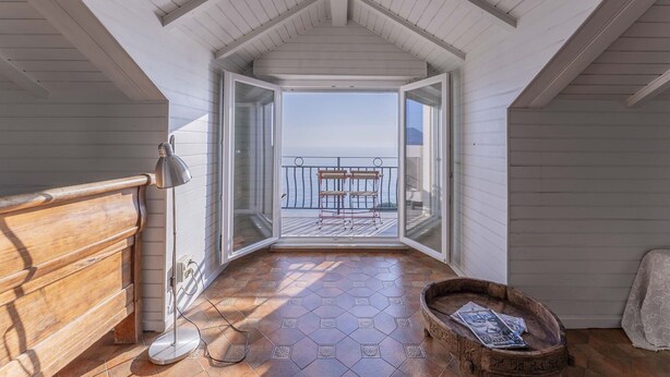 Um quarto estreito, com pé-alto e vista para o mar, um banco de madeira, um abajur e um porta-revistas.