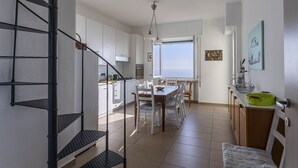 Uma cozinha moderna com armários brancos, uma área de jantar com mesa e cadeiras de madeira, uma escada com corrimão de metal e vista para o mar através de amplas janelas.